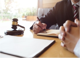 A man in a suit writes in a notebook while a gavel and blank papers rest on a wooden table.
