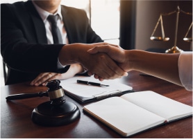 A lawyer in a suit shakes hands with a client over a desk with a gavel, notebook, and scales of justice in the background.