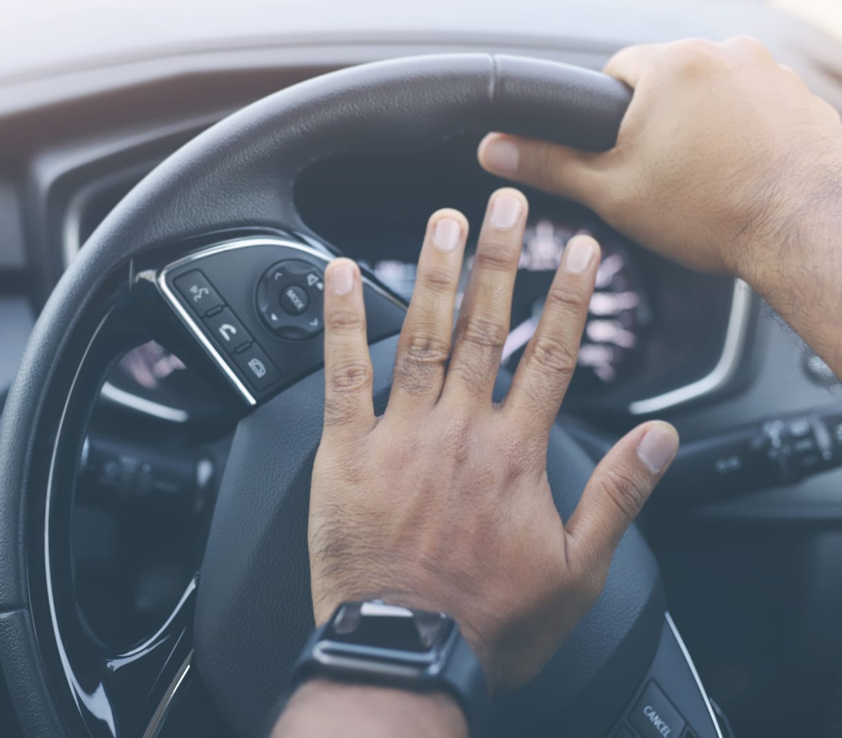 A close-up of a driver's hands on the steering wheel, one hand raised in a waving gesture while the other grips the wheel.